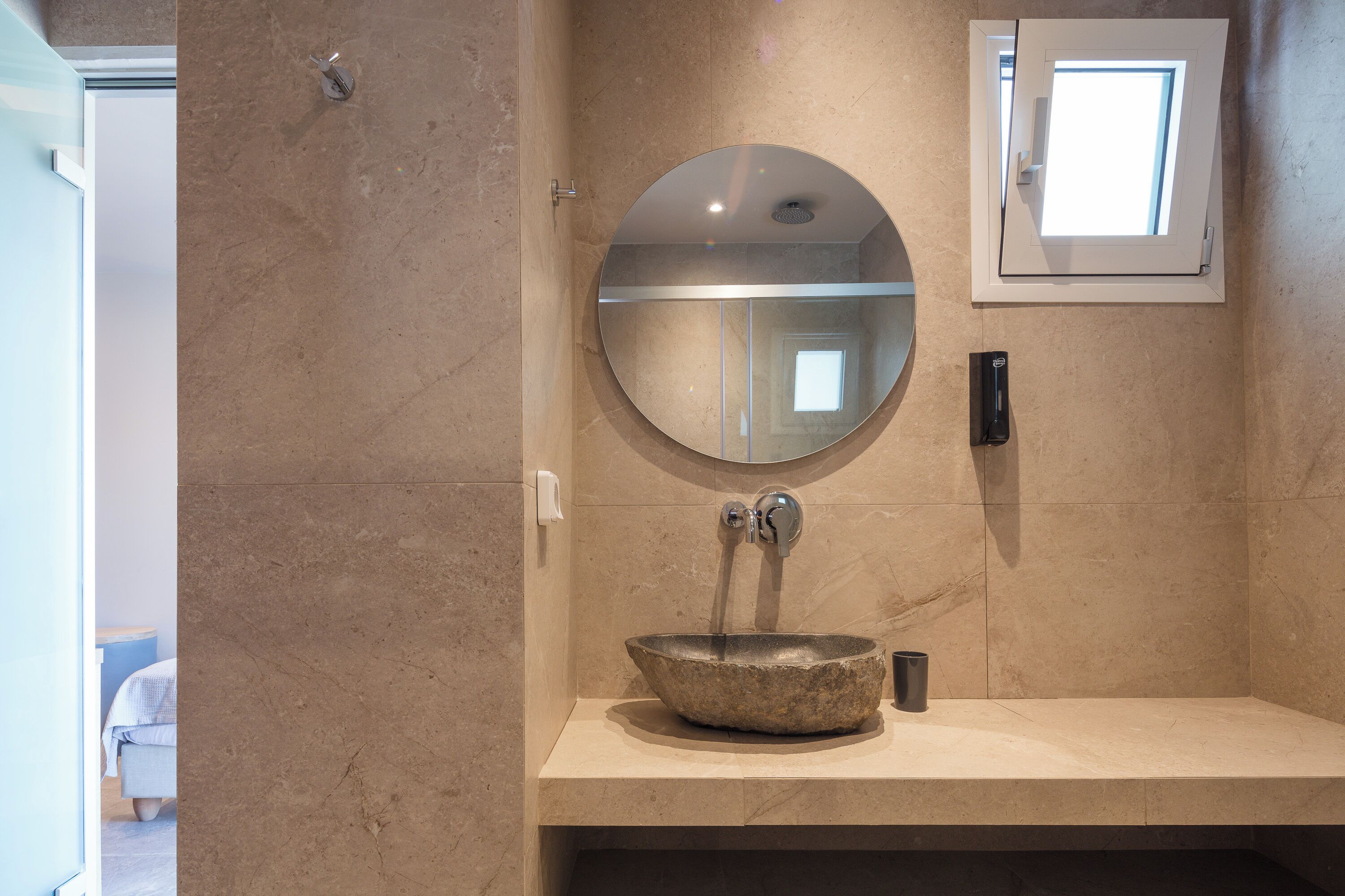 A bathroom with a round mirror, a stone sink, and a small window.