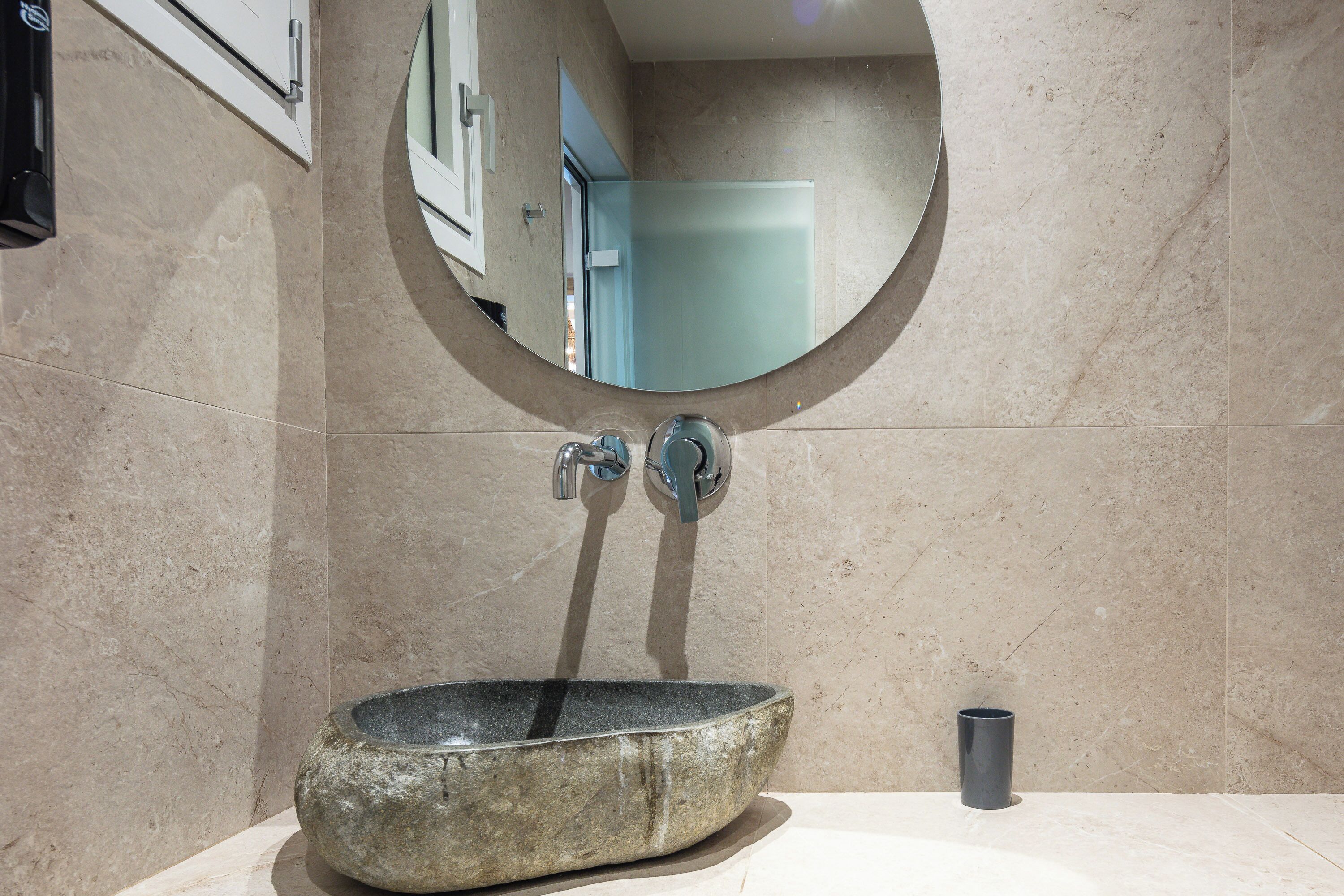 A bathroom with a stone sink, a round mirror, and a small cup on the counter.