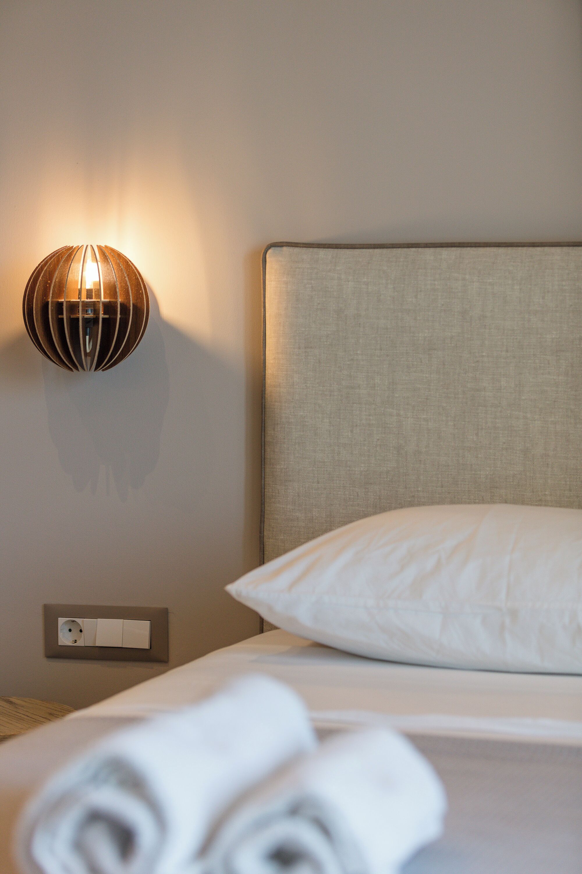 A neatly made bed with a white pillow and rolled towels, a wall-mounted light fixture, and a power outlet on the wall.
