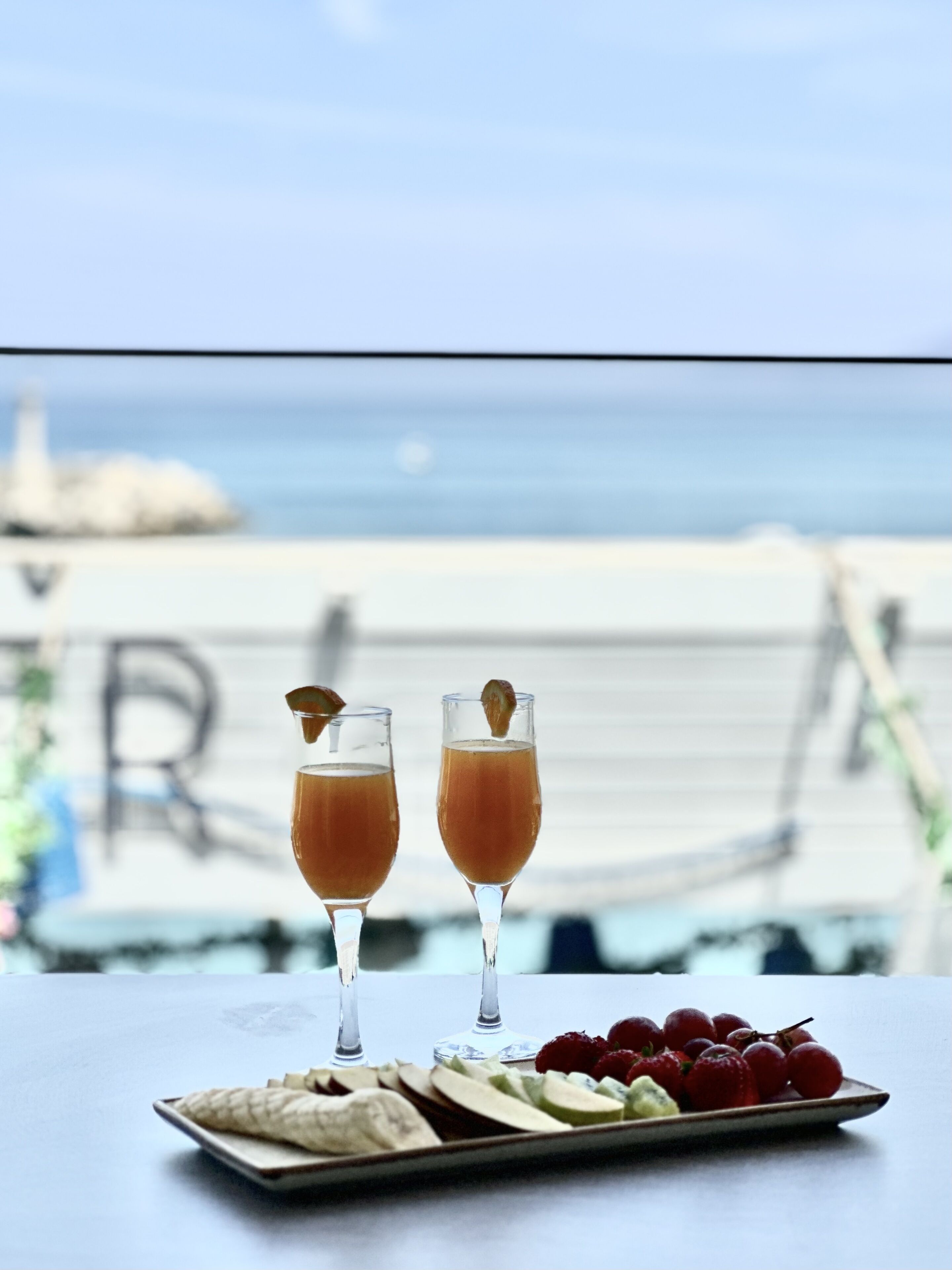 Two glasses of orange juice with garnishes on a table with a plate of fruit and crackers. A view of the beach and ocean is visible through a window.