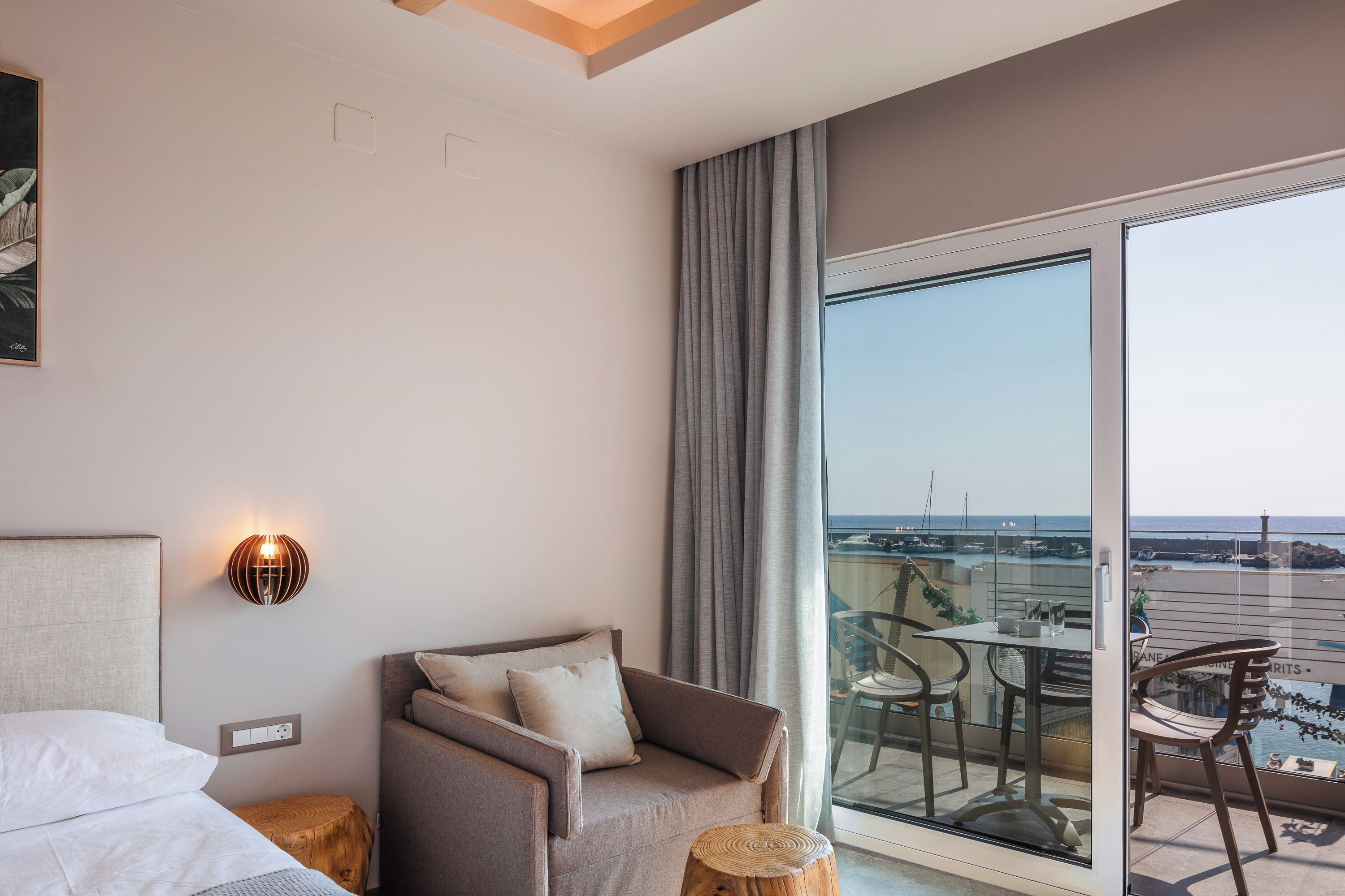Habitación de hotel con cama, sofá, taburete de madera, mesa con sillas y balcón con vistas al mar.