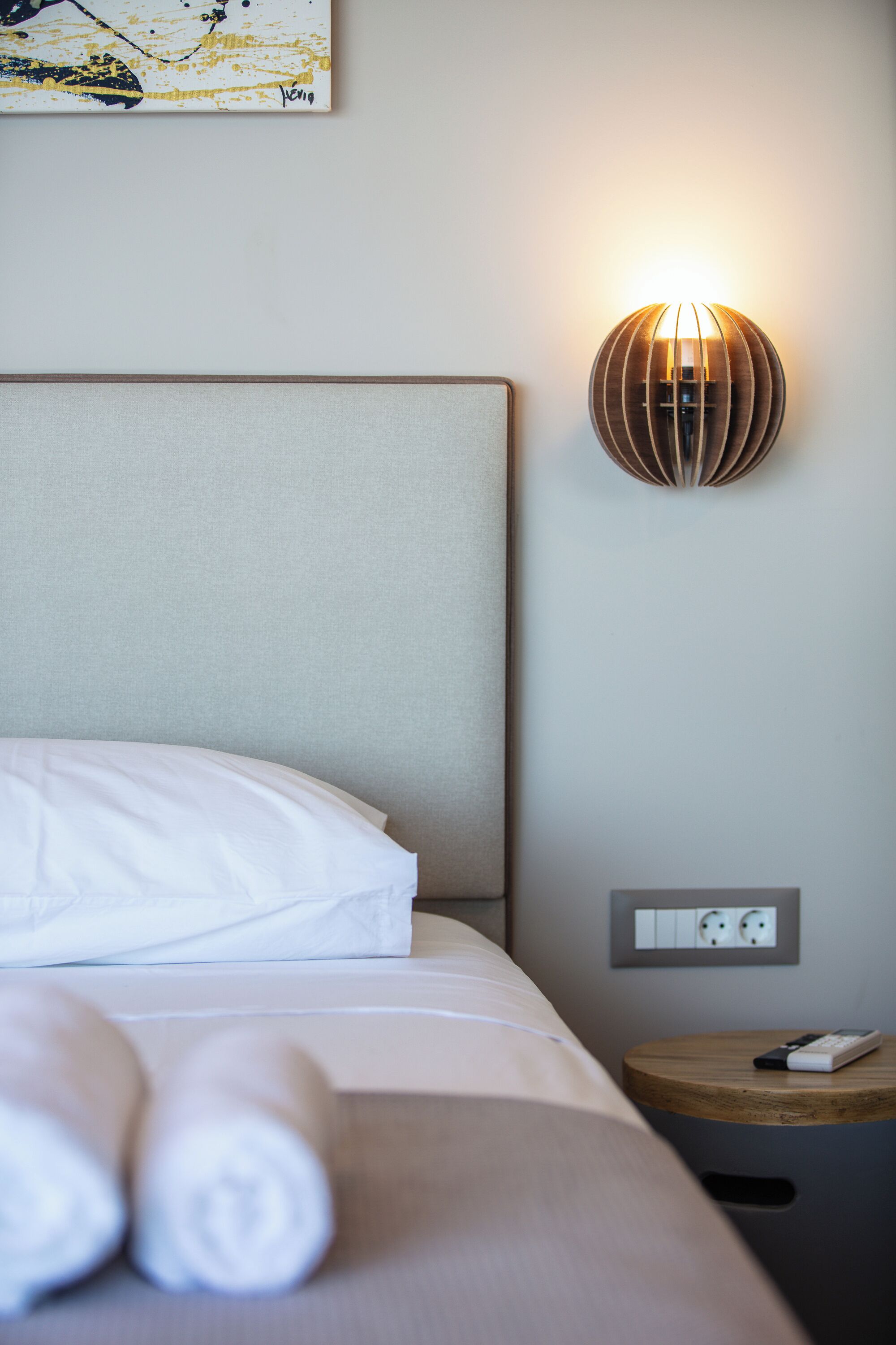 A neatly made bed with white linens and rolled towels, a wall-mounted light fixture, and a small wooden side table with a remote control.
