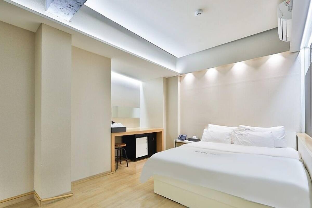 Quarto de hotel moderno com uma cama grande, uma escrivaninha com cadeira, um banquinho pequeno e um espelho na parede.