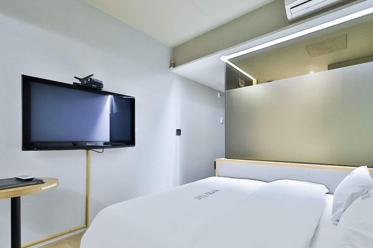 Quarto de hotel moderno com televisão de tela plana na parede, mesa de cabeceira com abajur e uma cama grande com roupa de cama branca.