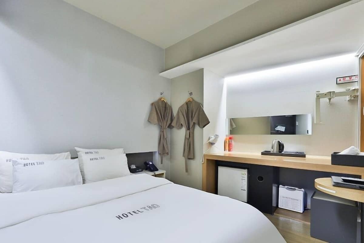 Quarto de hotel com cama, almofadas, escrivaninha com computador, espelho e um pequeno frigorífico.