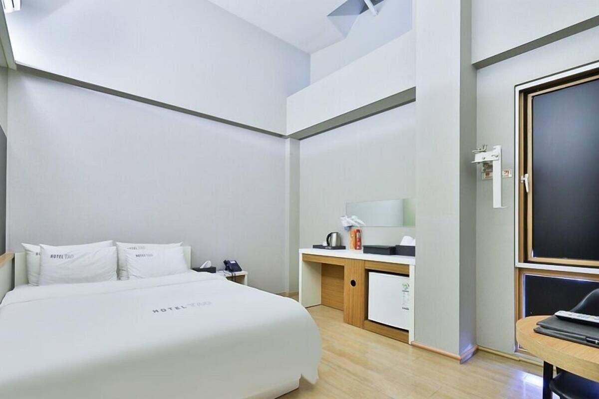 Quarto de hotel com cama, mesa de escritório com computador, pequeno espaço de cozinha e janela.