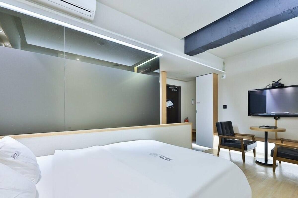 Quarto de hotel com cama, televisão, duas cadeiras e uma mesa pequena.