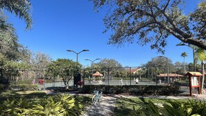 A tennis court surrounded by a fence, with a pathway leading to it. There are benches and a small kiosk nearby. - Vacation in Paradise, Naples, FL (Naples)