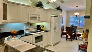 A modern kitchen with white cabinets, a microwave, stove, and refrigerator. There's a dining area with a table and chairs, and a ceiling fan. - Vacation in Paradise, Naples, FL (Naples)