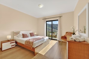 A modern bedroom with a large bed, bedside tables, a desk, and a view of a mountainous landscape through a sliding glass door. - Villa Bella - Mountain views & Outdoor living (Machico)