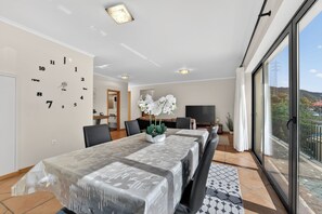 A dining area with a table covered in a patterned tablecloth, chairs, a clock on the wall, and a view of the outdoors through a sliding door. - Villa Bella - Mountain views & Outdoor living (Machico)