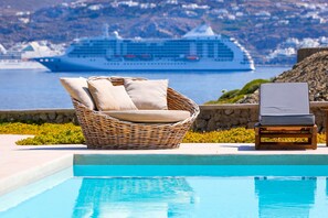 A poolside area with a wicker chair and a lounge chair, a cruise ship in the background, and a stone wall.