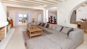 Un salon spacieux avec un grand canapé d’angle, une table basse en bois et une vue sur la mer grâce à de grandes fenêtres.