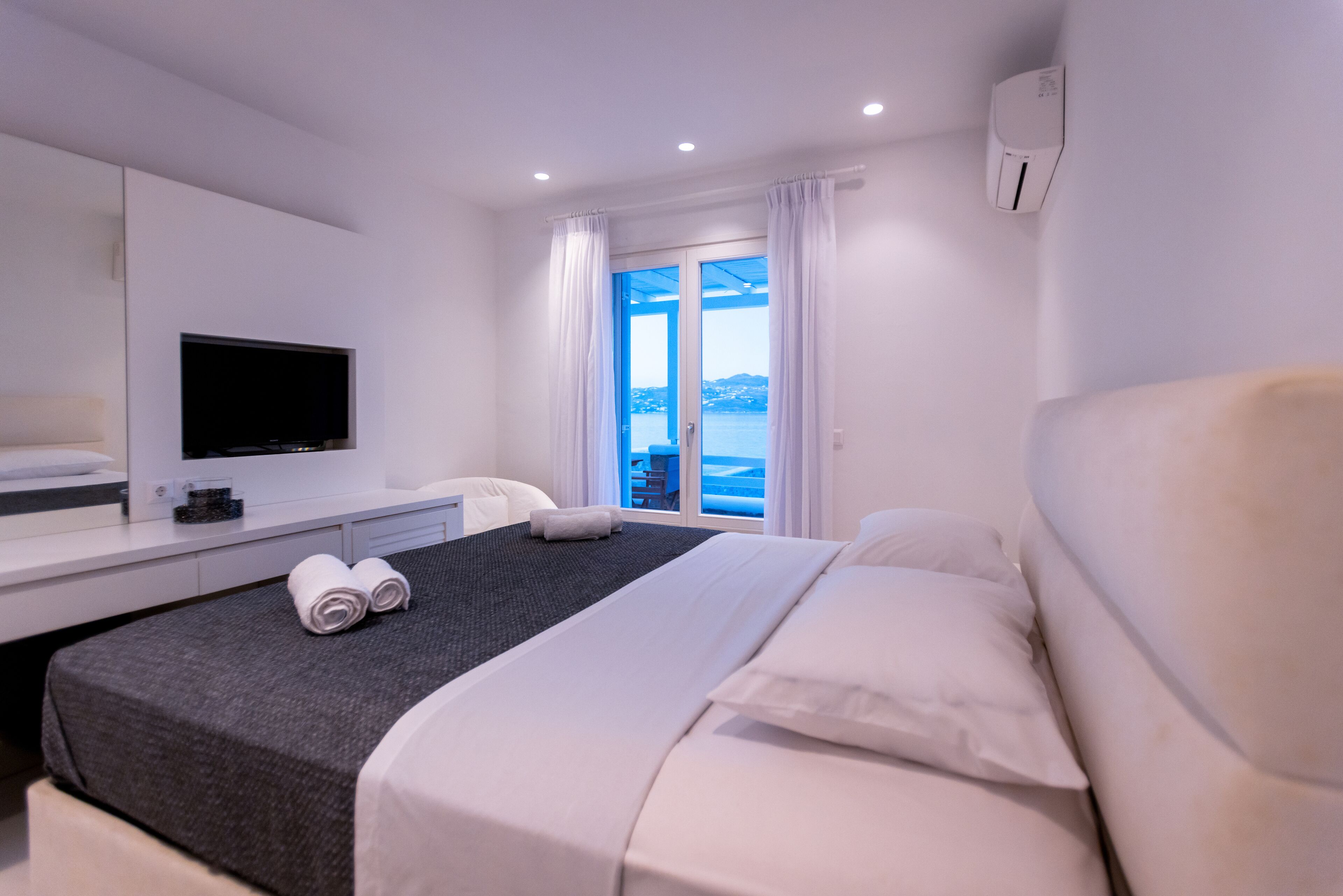 A modern hotel room with a large bed, a flat-screen TV, a desk, and a view of the sea through a glass door.