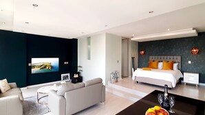 A modern hotel room with a large bed, a sofa, a TV, and a dining area with a bottle and glasses.