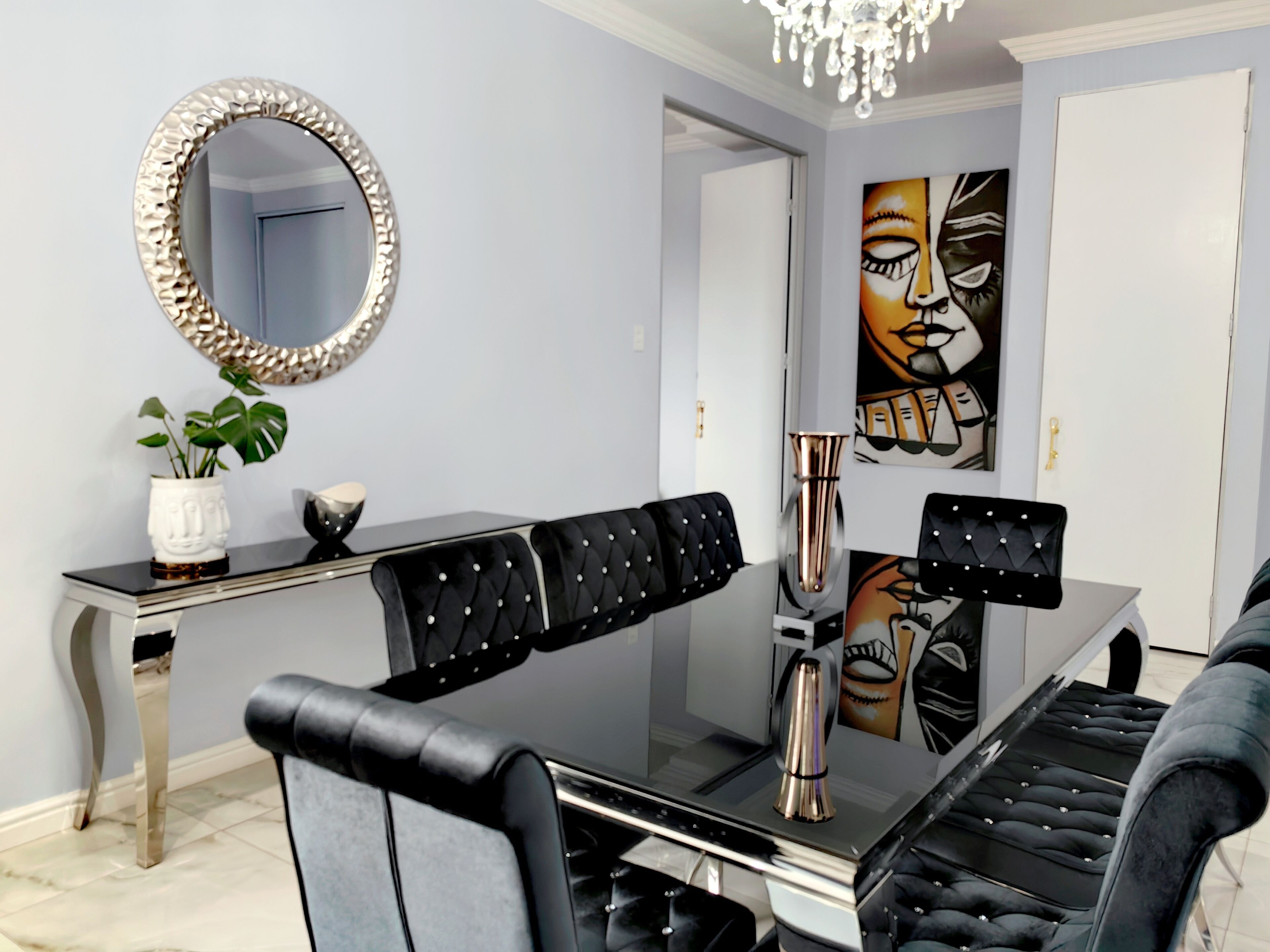 A modern dining area with a glass table, black upholstered chairs, a decorative mirror, and a chandelier.