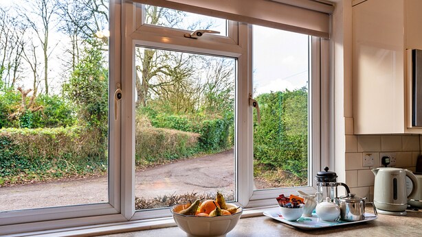 A kitchen with a large window, a bowl of fruit, a tray with a teapot and a kettle, and a view of a rural path.