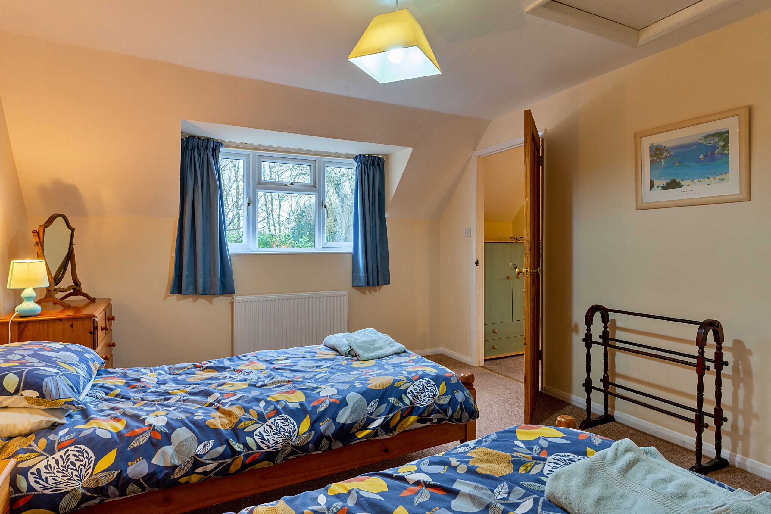 A bedroom with two beds, a window with curtains, a radiator, a wooden dresser, a lamp, and a framed picture on the wall.