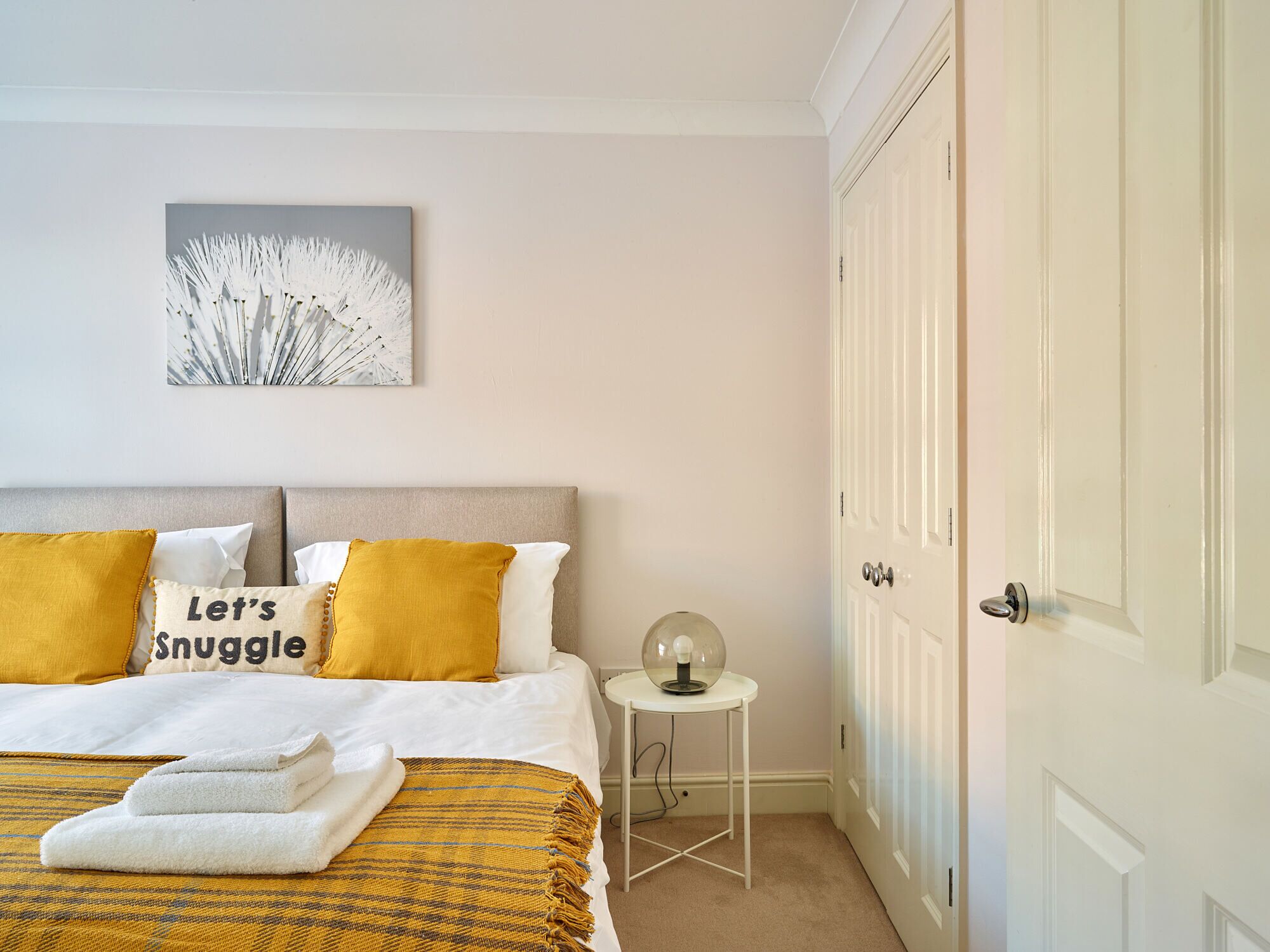 Un dormitorio ordenado con cama, almohadas, una mesita de noche con lámpara y una puerta.