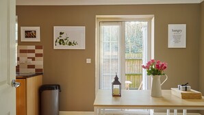 A kitchen with a window, a trash can, a table with a vase of flowers, and a framed picture on the wall.