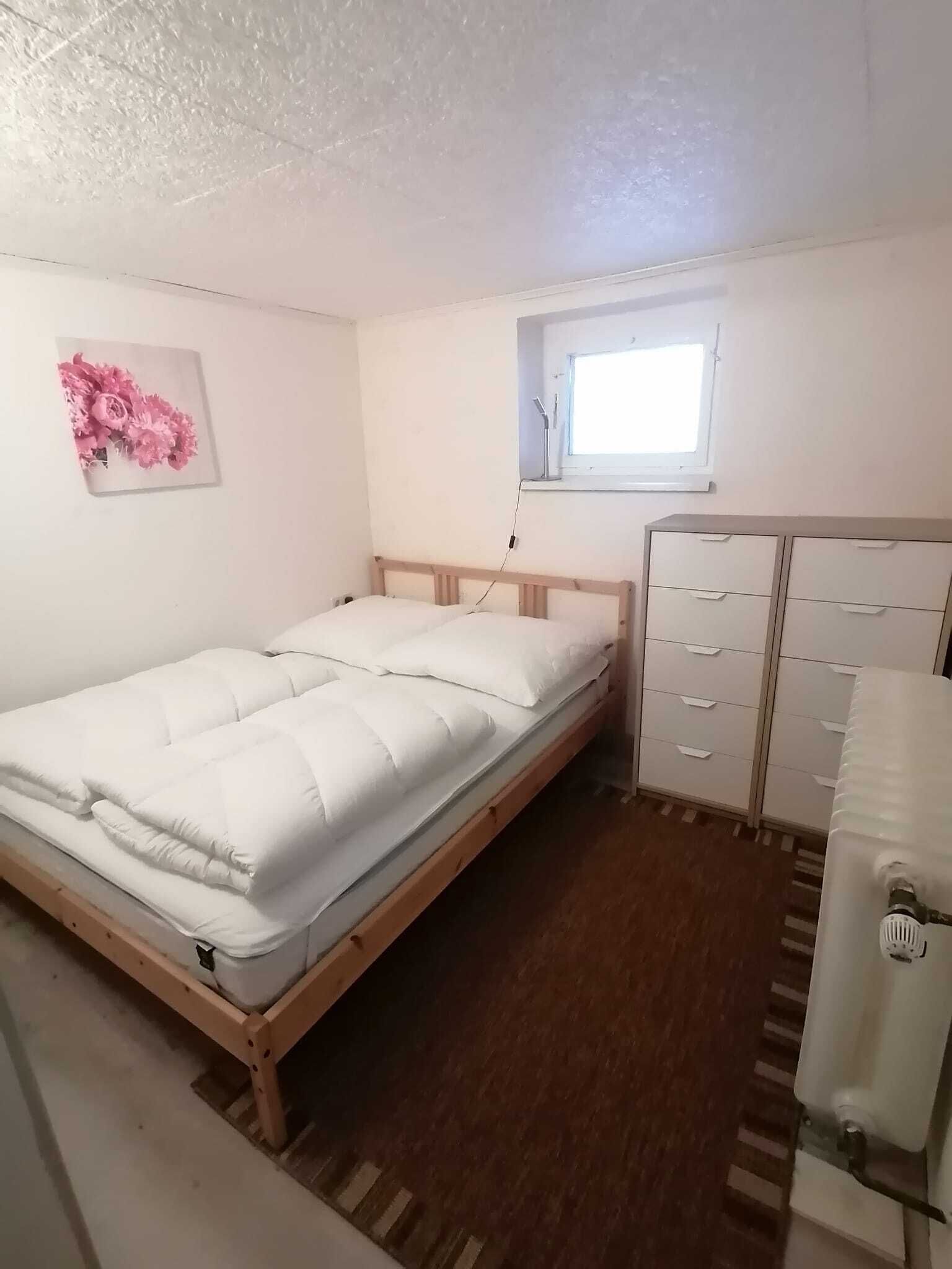 Une petite chambre bien éclairée, avec un lit simple, une fenêtre, un radiateur et une commode.