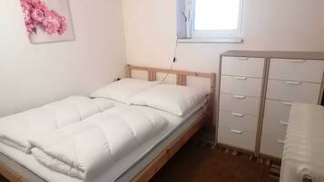 Une petite chambre bien éclairée, avec un lit simple, une fenêtre, un radiateur et une commode.