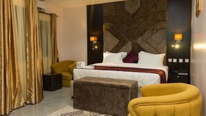 A modern hotel room with a large bed, a sofa, a small table, and a decorative wall panel. - Iris Gold Suite (Ibadan)