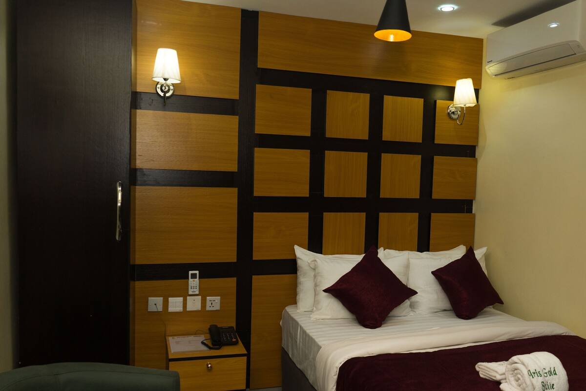 A hotel room with a bed, bedside table, lamp, and an air conditioning unit.