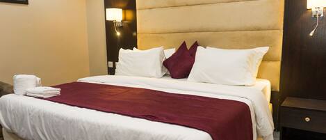 A hotel room with a large bed, white linens, a red throw blanket, and a nightstand with a lamp.