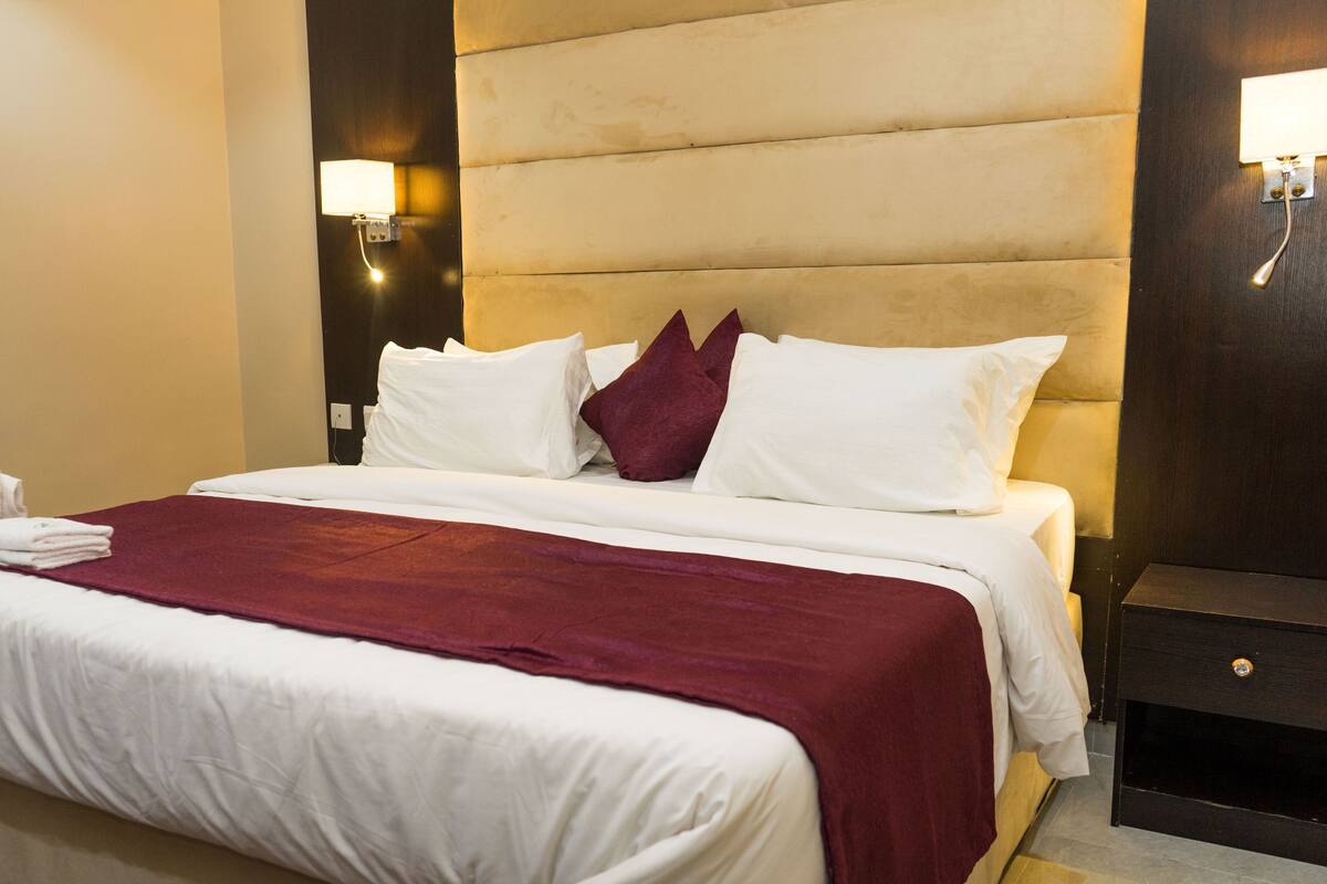 A hotel room with a large bed, white linens, a red throw blanket, and a nightstand with a lamp.