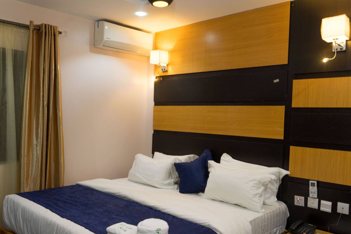 A hotel room with a bed, curtains, an air conditioning unit, a wall-mounted light, and a phone on the nightstand.