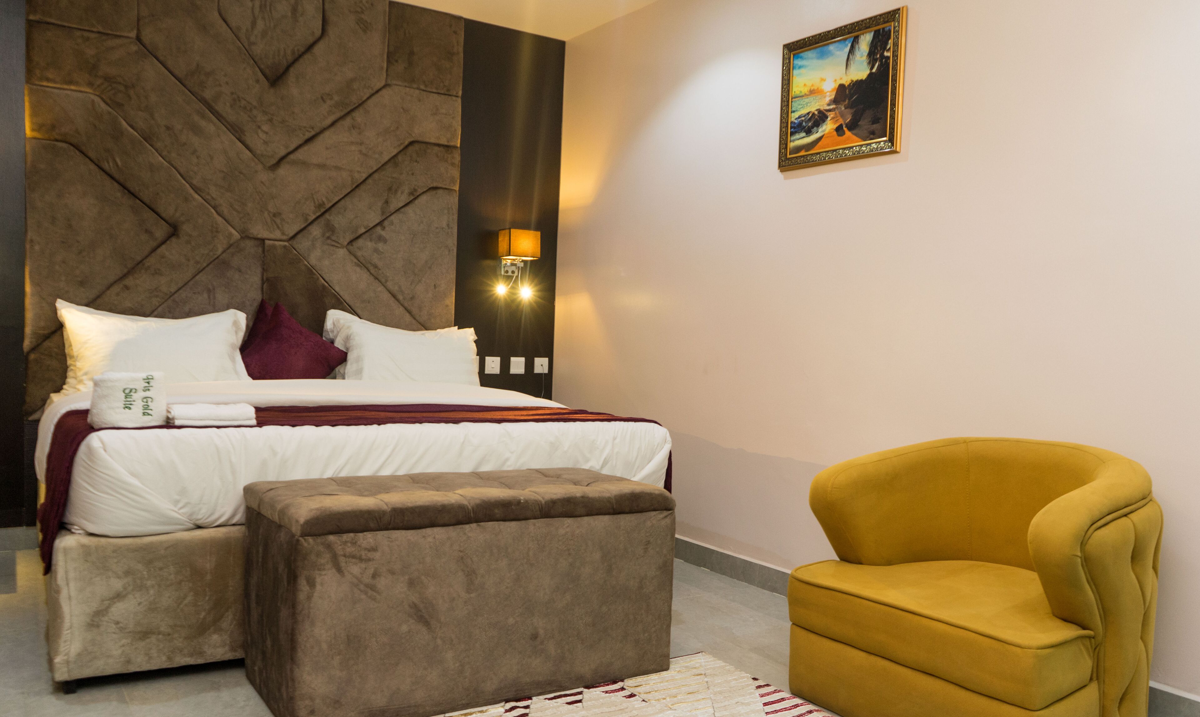 A modern bedroom with a bed, a headboard, a wall-mounted light, a painting, and a yellow armchair.