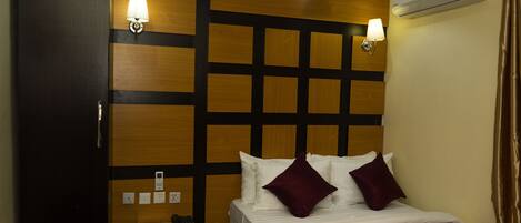 A hotel room with a bed, bedside table, lamp, and an air conditioning unit.
