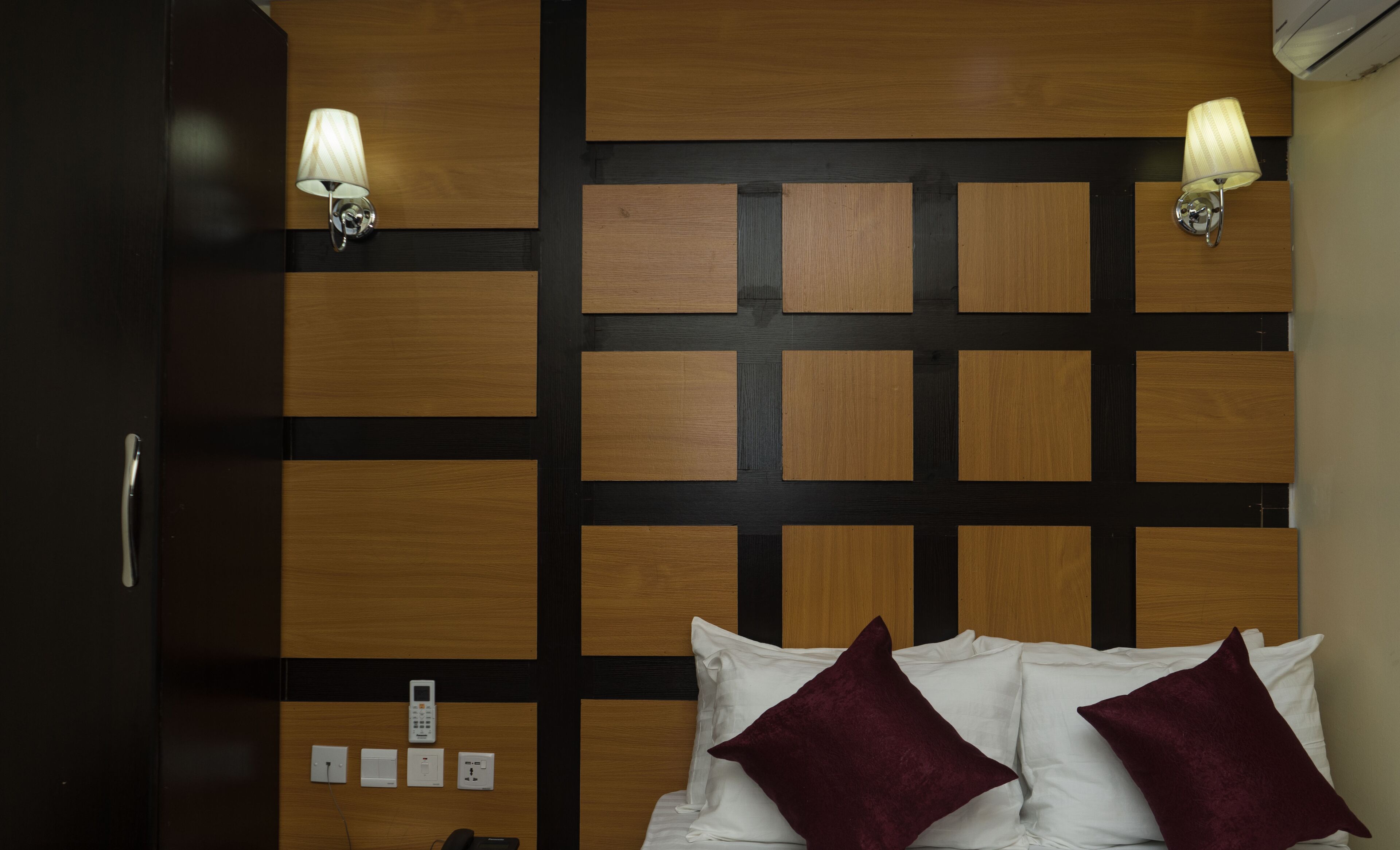 A hotel room with a wooden headboard, two wall-mounted lamps, a bed with white linens and two burgundy pillows, and a bedside table with a phone and a power outlet.