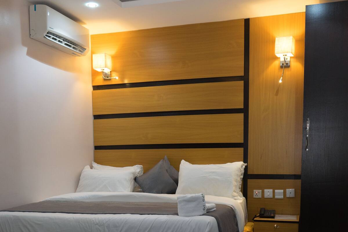A modern hotel room with a large bed, bedside tables, an air conditioning unit, and a desk.