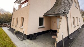 Une maison à deux étages, dotée d’un toit en pente, d’un porche couvert et d’un petit espace extérieur aménagé pour s’asseoir.