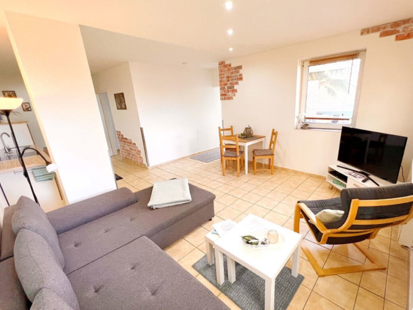 A living room with a grey sofa, a small white table, a wooden chair, a TV, and a brick accent wall.