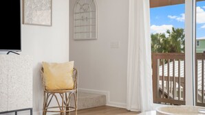 A living room with a television, a wicker chair with a cushion, a decorative wall piece, and a view of the outdoors through a glass door.