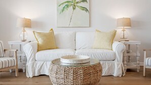 A white sofa with yellow cushions, two side tables with lamps, and a woven coffee table.