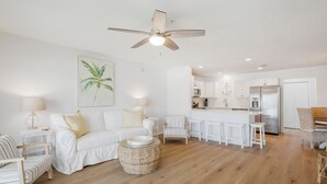 A spacious living room with a white sofa, a dining area with a bar, and a kitchen with white cabinets and a stainless steel refrigerator.