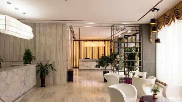 Um hall de hotel moderno com piso de mármore, cadeiras brancas, balcão de recepção e uma estante repleta de plantas.