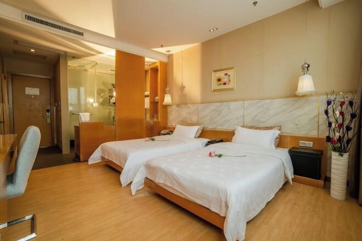 A hotel room with two beds, a desk, a chair, a nightstand, a vase with flowers, and a framed picture on the wall.