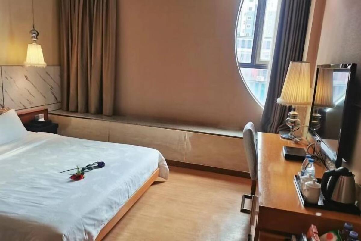 Et hotelværelse med seng, skrivebord, lampe, fjernsyn og et vindue med gardiner.