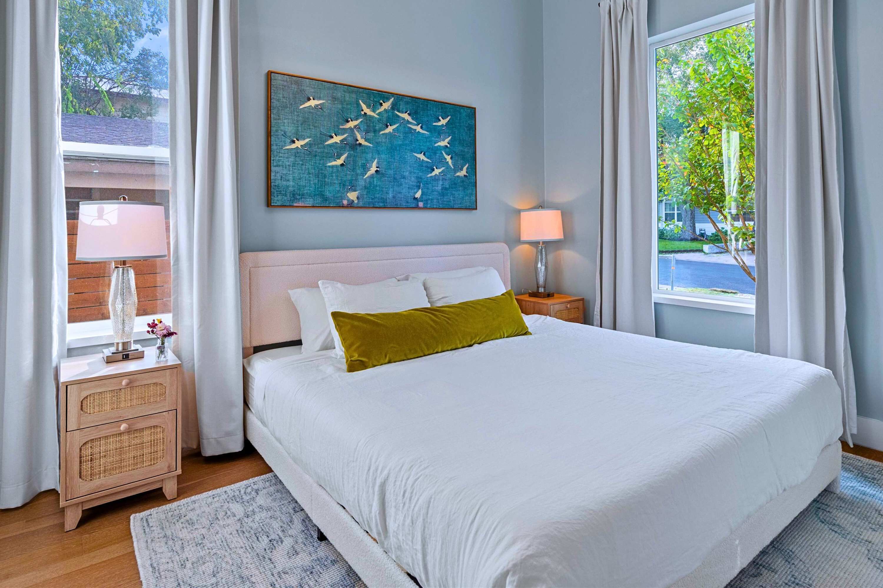 A bedroom with a large bed, two bedside lamps, a nightstand, a decorative painting, and a view of the outdoors.