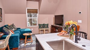 A cozy living room with a teal sofa, a small round table, and a kitchen area with a sink and a vase of flowers. - East Nashville Guesthouse close to everything & Pet-Friendly! (Nashville)