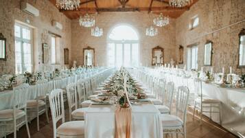 A long banquet hall with white chairs and tables, chandeliers, and large windows.