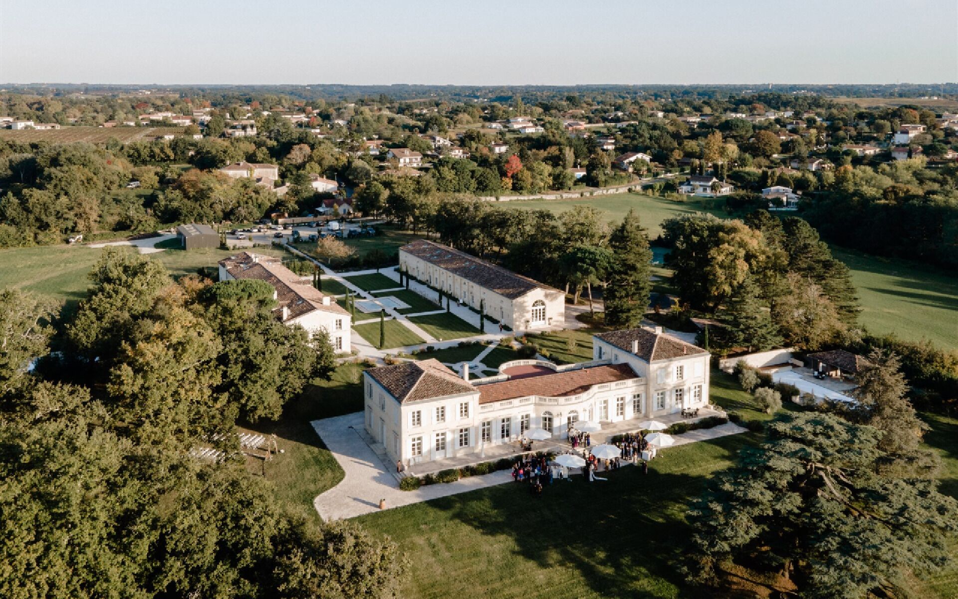 Um edifício grande, branco e de vários andares, com fachada simétrica, cercado por jardins e árvores bem cuidados.