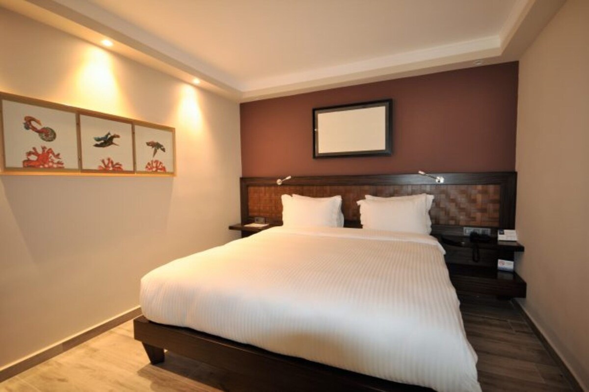 Ein Hotelzimmer mit einem großen Bett, einem Nachttisch, einem wandmontierten Fernseher und gerahmten Kunstwerken an der Wand.