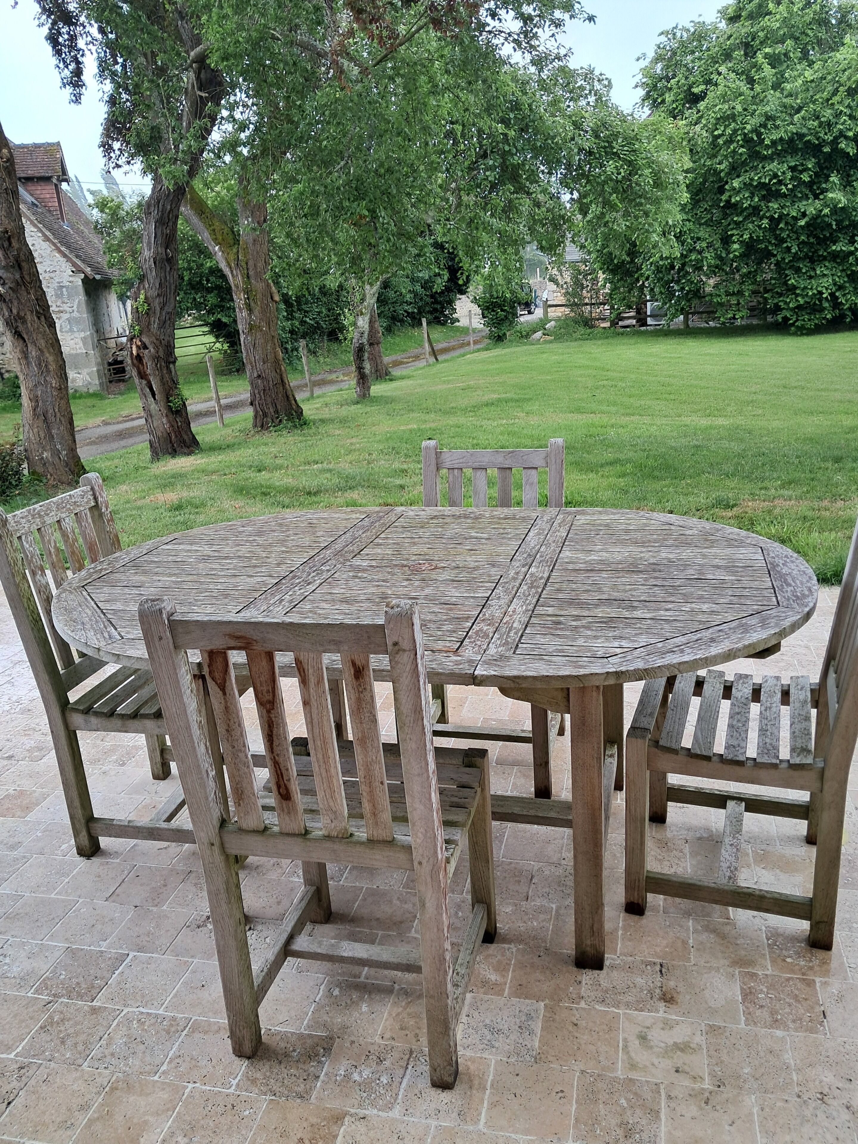 Una mesa redonda de madera con sillas, sobre baldosas de piedra, rodeada de árboles y un área de césped.