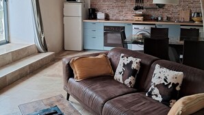 A cozy living room with a brown leather sofa, a wooden coffee table, and a kitchen area with a brick backsplash, a stove, and a window.